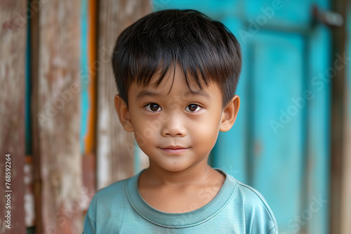 A young nepalese boy with dark hair and a white shirt is smiling at the camera. The boy's eyes are closed, and he has a slight smirk on his face. The scene is set outdoors photo