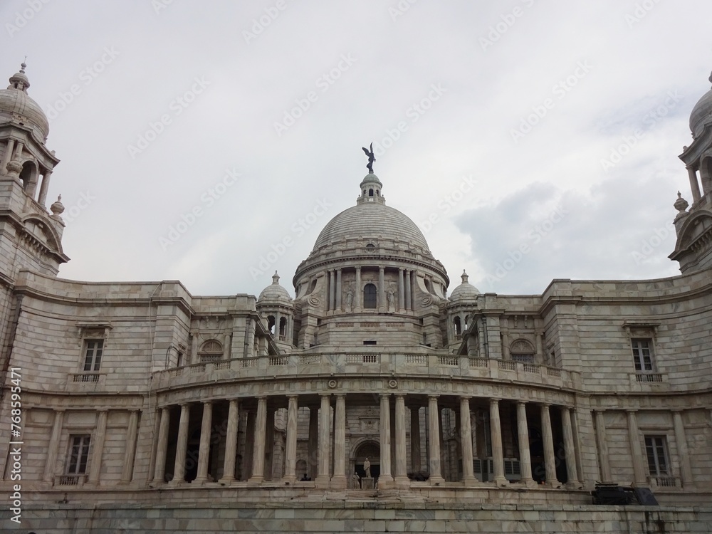 The grand Victoria Memorial Hall stands under a dramatic cloud-filled sky, showing off its exquisite colonial architecture with its towering domes and intricate stonework.