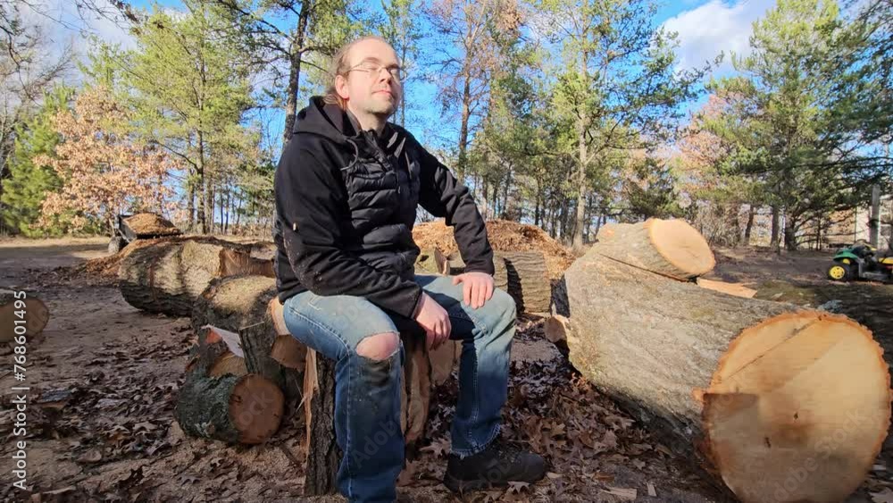 Man in black jacket wearing glasses and blue jeans with holes sitting on stump or block of wood taking break after processing oak logs into firewood. Man in black jacket wearing glasses and blue jeans with holes sitting on stump or block of wood taking break after processing oak logs into firewood.