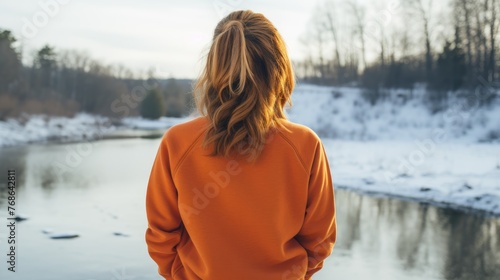 View from the back of a girl in plain orange crewneck sweatshirt mockup on a winter background with icicles and a frozen lake behind