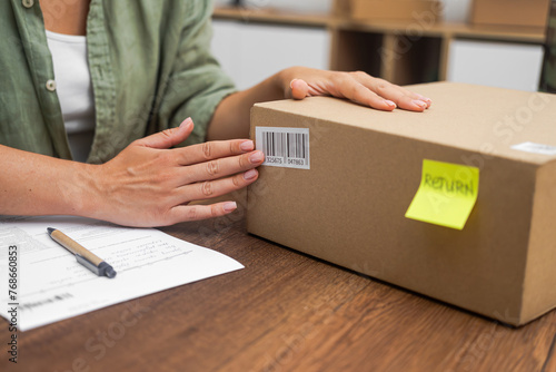A female shopper affixes a Return sticker, featuring a yellow label with a barcode, onto a cardboard box, initiating the online shopping refund process. 