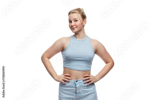 A smiling young girl stands with her hands on her belt. A cute blonde teenager with freckles on her face in a blue top and jeans. Positivity, optimism and charm. Isolated on a white background.