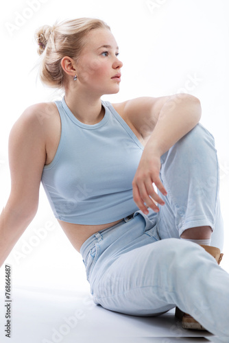 A young teenage girl sits and looks to the side. Cute blonde teenager with freckles on her face wearing a blue top and jeans. White background. Close-up.