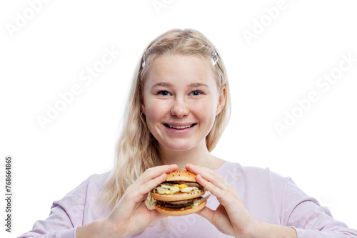 Smiling young girl with a big hamburger in her hands. Cute blonde teenager with freckles in a pink sweater. Delicious garbage food. Isolated on a white background.