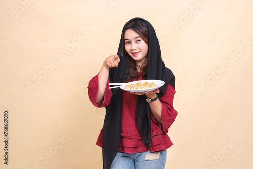 Beautiful mature indonesia woman wearing a red hijab smiling, pointing at the camera and carrying a plate containing chopsticks and dimsum (Chinese food). used for food, health and lifestyle content