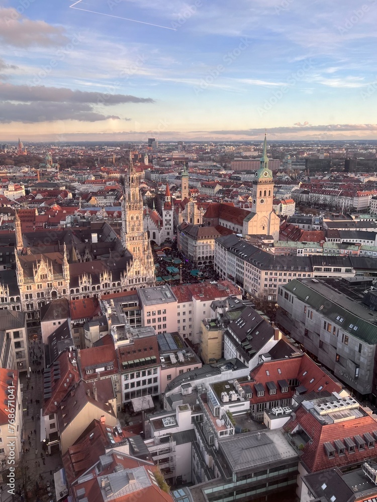 Fototapeta premium A breathtaking aerial view of Munich's cityscape at sunset, showcasing the iconic gothic architecture of the New Town Hall and the St. Peter's Church spires against a backdrop of the distant Alps