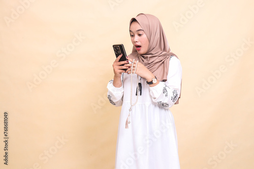 a young Asian Muslim woman wearing a hijab is shocked holding prayer beads and chatting via smartphone gadget. for advertising, lifestyle, banners and Ramadan