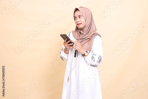 A young Asian Muslim woman wearing a hijab is thinking about holding prayer beads and WhatsApp via smartphone gadget. for advertising, lifestyle, banners and Ramadan