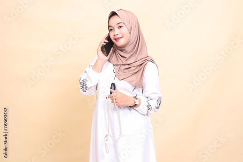 A young Asian Muslim woman wearing a hijab smiles candidly, holding prayer beads and calling someone. for advertising, lifestyle, banners and Ramadan