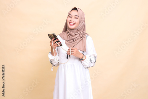 a young Asian Muslim woman wearing a hijab smiles cheerfully holding prayer beads and carrying a smartphone gadget. for advertising, lifestyle, banners and Ramadan