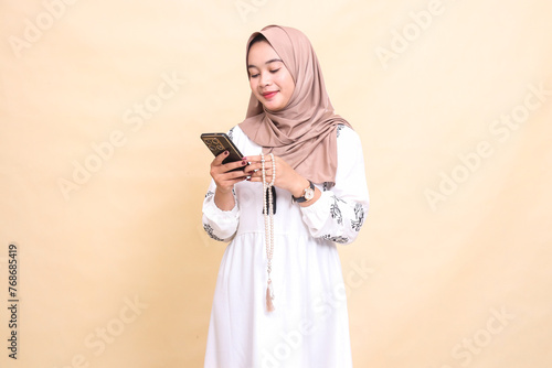 a young Asian Muslim girl wearing a hijab smiles holding prayer beads and chatting via cellphone gadget. for advertising, lifestyle, banners and Ramadan