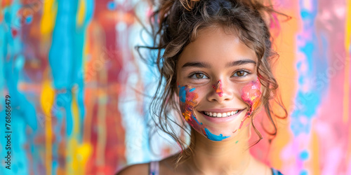 Delightful young girl with curly hair and face smeared with vibrant paint colors, smiling brightly in front of a colorful backdrop