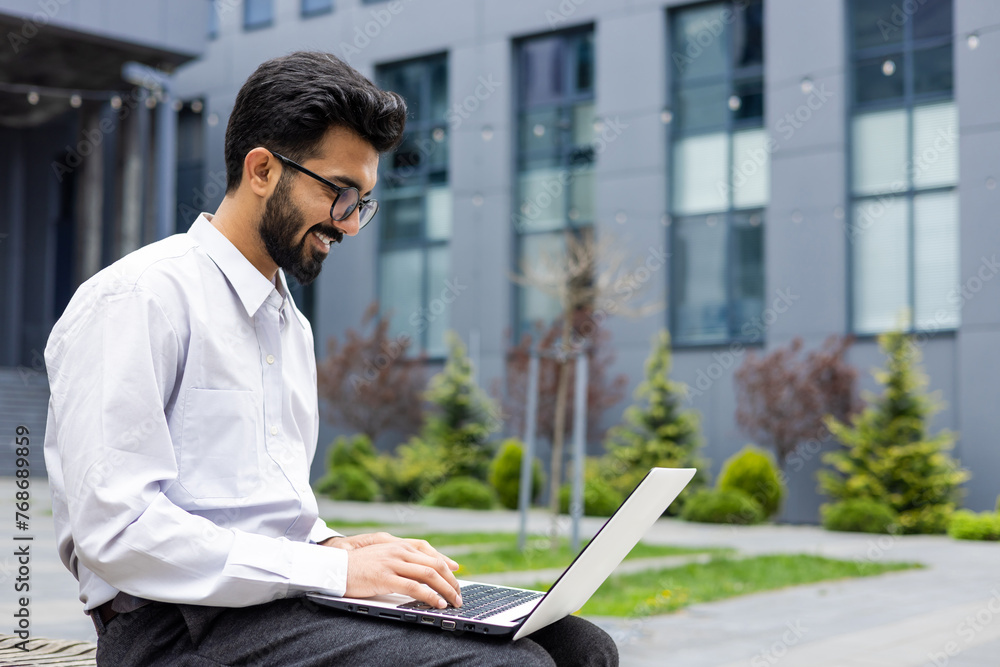 Smiling young Indian male office worker, student sitting outside business building and working, studying online at laptop. Smiling young Indian male office worker, student sitting outside business building and working, studying online at laptop.