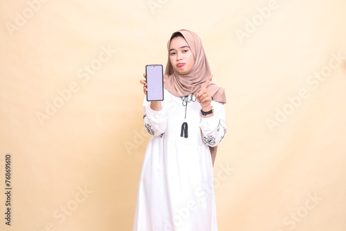 An indonesia Muslim woman wearing a hijab frowns showing a cellphone (gadget) screen pointing up to create sales content. for advertising, technology, Eid and Ramadan