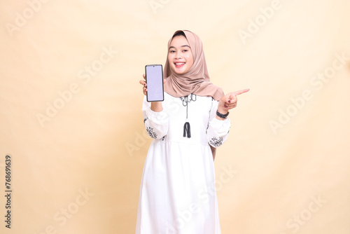 an adult indonesia Muslim woman wearing a hijab with a cheerful smile displays a cellphone screen (gadget) pointing left to create sales content. for advertising, technology, Eid and Ramadan