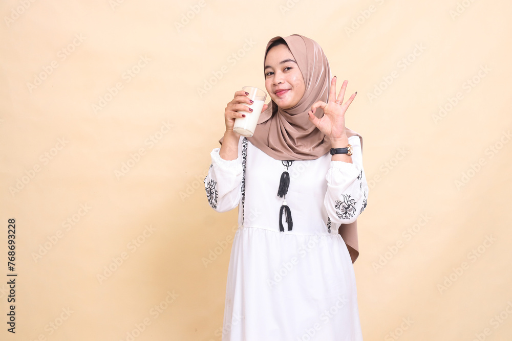 a beautiful Asian Muslim woman wearing a hijab, smiling with a good gesture and bringing a glass of white milk for drinks. used for advertising, health, Eid and Ramadan a beautiful Asian Muslim woman wearing a hijab, smiling with a good gesture and bringing a glass of white milk for drinks. used for advertising, health, Eid and Ramadan