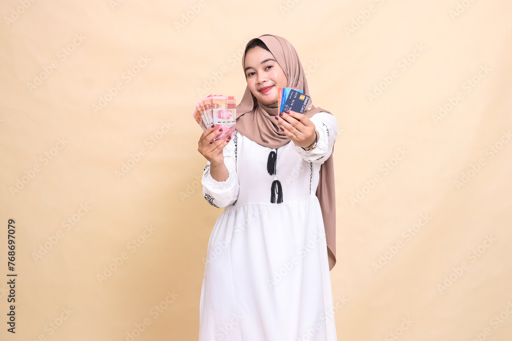 A beautiful Asian Muslim woman wearing a hijab with a smiling expression carries rupiah money and advances a debit credit card for shopping payments. used for advertising, technology, Eid and Ramadan A beautiful Asian Muslim woman wearing a hijab with a smiling expression carries rupiah money and advances a debit credit card for shopping payments. used for advertising, technology, Eid and Ramadan