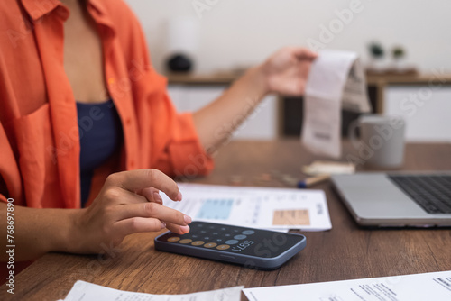 Expense Management: Seated at a desk with a laptop, a woman uses her smartphone calculator to compute household finances