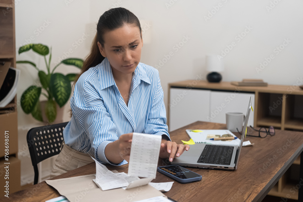 A lady, at home, holds a bill, using her laptop for online payments. She calculates taxes, reviews accounts, and loan rates. A lady, at home, holds a bill, using her laptop for online payments. She calculates taxes, reviews accounts, and loan rates.