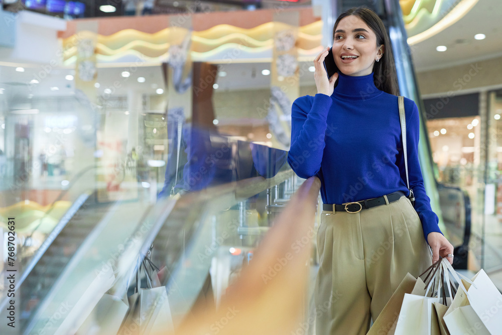 Front view portrait of smiling young woman speaking on smartphone in shopping mall and holding paper bags copy space
