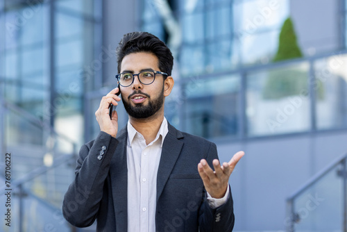Canvas Print Close-up photo of a busy young Indian businessman man in a suit and glasses standing near an office center on the street, talking seriously on the phone and gesturing with his hand