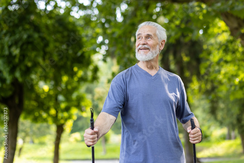 Portrait of an older gray-haired man standing in a park with trekking poles, doing Nordic walking. He looks away with a smile.