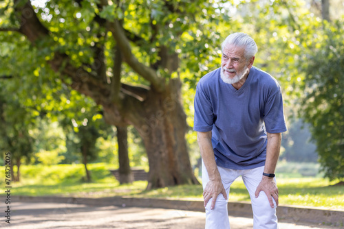 Tired senior gray-haired man is standing bent over in the park and resting after an active walk, run and workout.