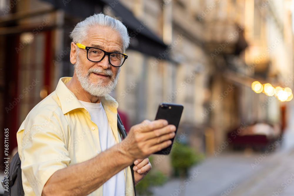 Portrait of a senior gray-haired smiling man walking and traveling in the city with a rucksack, holding a phone in his hand and looking happily at the camera. Portrait of a senior gray-haired smiling man walking and traveling in the city with a rucksack, holding a phone in his hand and looking happily at the camera.