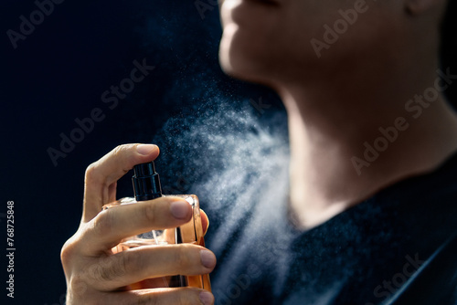 Fragrance spray. Man and perfume. Applying cologne. Scent water. Skin care, beauty product and male cosmetics concept. Holding elegant smell bottle in hand. Dark blue background. Young gentleman. photo