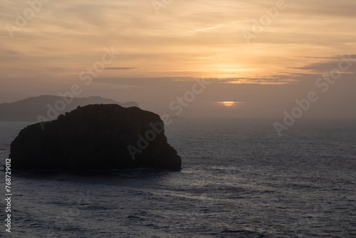 a peaceful sunset behind an imposing rock formation amidst the ocean, evoking a sense of tranquility and natural beauty