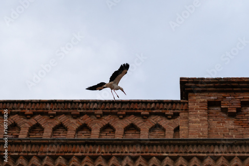Stork is flying over a building with clouds in the background.
