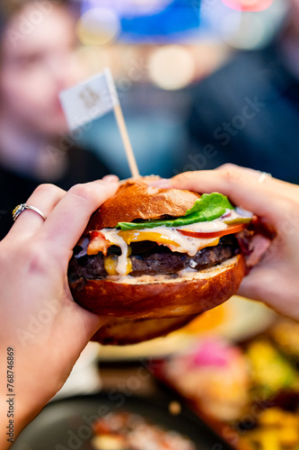 person’s hands hold a juicy burger with melted cheese, lettuce, and sauce. The glossy bun is topped with a small flag-like stick. background is blurred, and the person’s face is pixelated for privacy