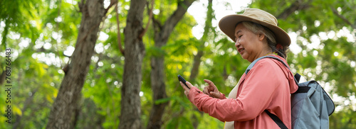 Portrait of Asian mature woman holding smartphone and backpack behind back, an Asia active senior woman enjoying nature in park. Standing on a trail in a forest outdoors. Enjoying active travel trip