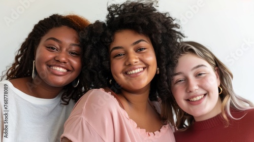 Three smiling young women with curly hair wearing casual clothing standing close together posing for a photo with a white background.