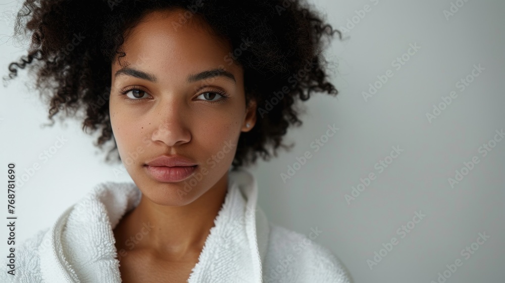 A young woman with curly hair and a soft expression wearing a white robe looking directly at the camera. A young woman with curly hair and a soft expression wearing a white robe looking directly at the camera.