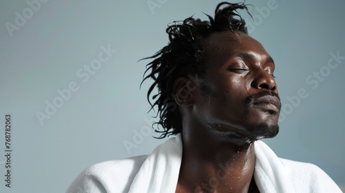 A man with a beard and dreadlocks eyes closed enjoying a relaxing moment with water dripping from his face wrapped in a white towel against a blue background.