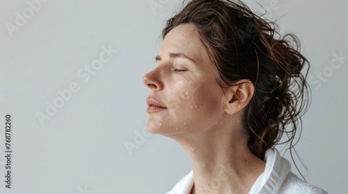 A woman with closed eyes enjoying a moment of relaxation with water droplets on her face set against a soft light background.