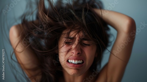 A woman with long hair clenching her teeth in distress with her hands on her head against a blue background.