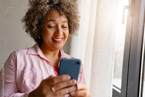 A mature woman of mixed race standing near large window, holding a smartphone and smiling. Mobile phone apps for older people