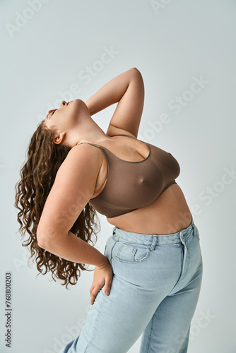 plus size young woman in brown bra and blue jeans with curly hair putting head to behind with hand
