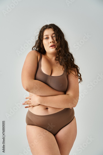 alluring plus size young woman in underwear with curly brown hair hugging herself on grey background