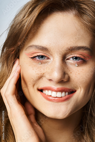 Closeup beauty portrait of smiling girl with tear face jewels, peach makeup and freckles on grey