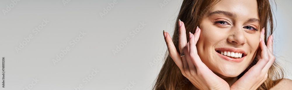 Tender young woman with perfect skin smiling at camera touching face, on grey background, banner Tender young woman with perfect skin smiling at camera touching face, on grey background, banner