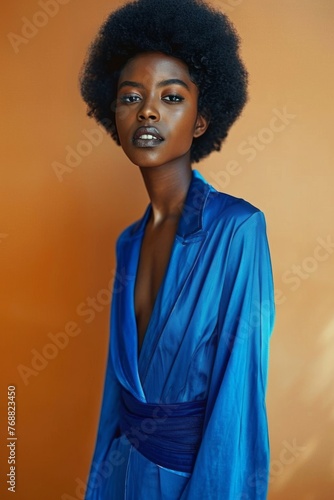 Half-length portrait of young African American woman in blue silk suit against orange studio background. Beautiful sexy black model with afro hairstyle looks seductively at camera.