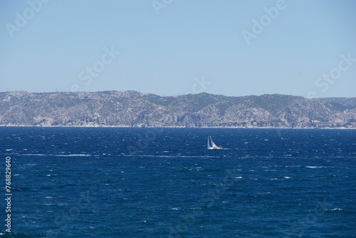 Sailboat gliding across a sparkling blue sea with mountains and a blue sky in the background © Wirestock