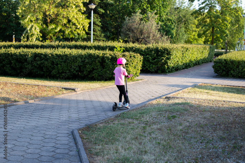 A beautiful girl in a pink protective outfit rides a scooter through the streets of the city in summer. Safe cycling and scootering without injury.
