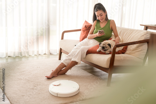 Young asian woman relaxing with pug dog in living room while Robotic vacuum cleaner working.