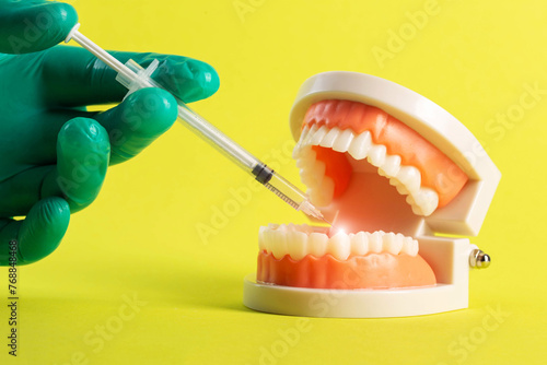 The hand of a dentist doctor in a green medical glove makes an anesthetic injection into a mock-up of a dental jaw on a yellow background. Concept of anesthesia and freezing during dental treatment.  photo