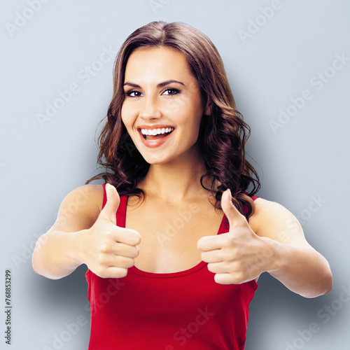 Portrait of happy smiling woman wear casual clothing, show thumbs up gesture, against grey wall background. Girl in red dress. Brunette excited model at studio. Square image.