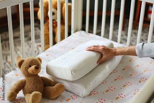 person placing an orthopedic pillow in a baby crib, teddy bear beside